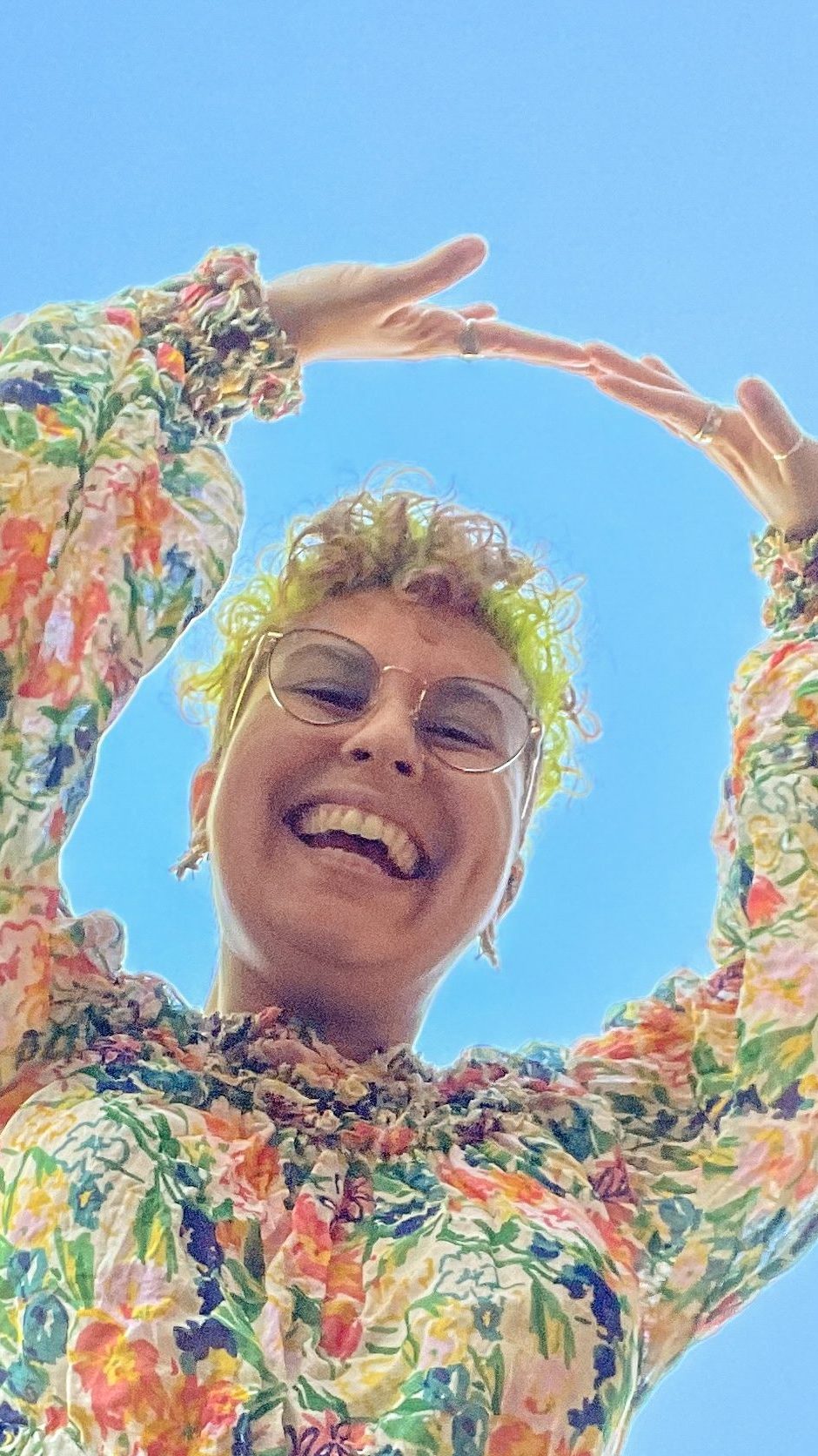 an image of Ela in a colorful top and jeans against a blue sky. Her hands are up above her head and there's a big grin on her face.